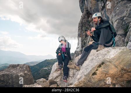 Konzept: Abenteuer. Paar Kletterer mit Helm und Geschirr. Auf einem Felsen sitzend. Mit dem Smartphone und Blick auf den Horizont. Über Stockfoto