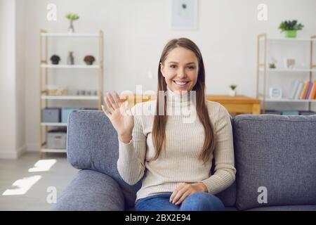 Junge Frau, die die Kamera anschaut, während sie online ihre Hand winkt, während sie mit einem Laptop mit einer Telefonverbindung mit einem virtuellen Mann im Zimmer ist. Stockfoto