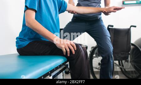 Physiotherapeut, der mit behinderten Menschen im Rollstuhl auf einem Therapietisch trainiert. Stockfoto