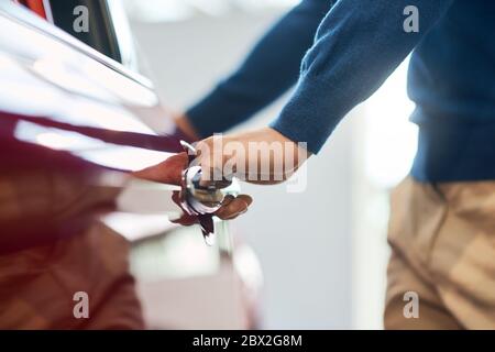 Hand des männlichen unerkennbaren Geschäftsmann öffnen und halten rote Autotür. Nahaufnahme des jungen kaukasischen männlichen Fahrer in lässigem Outfit öffnen Tür des Automobils in Autohaus. Stockfoto