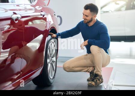 Schöner lächelnder bärtiger Mann, der sich hinsetzt, berührt und auf das Rad des roten Autos im Autohaus schaut. Seitenansicht des glücklichen Nachwuchses vor dem erfolgreichen Kauf. Stockfoto
