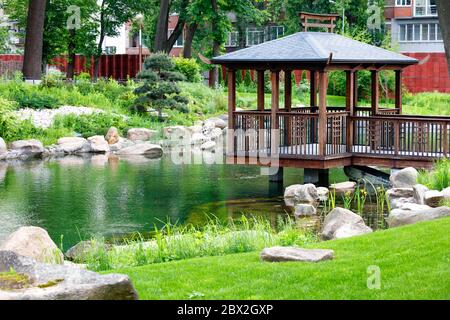 Holzdurchbrochene Laube auf Stelzen über dem Wasser eines Waldsees, umgeben von einem schönen Park mit Landschaftsgestaltung. Stockfoto