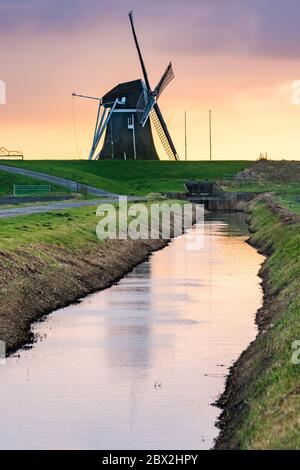 Eemshaven, Niederlande - 10. Januar 2020. Alte Windmühle hinter dem Wasserkanal bei Sonnenuntergang Stockfoto