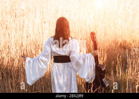 Rückansicht von schlanken jungen Frau mit roten Haaren in einem weißen mittelalterlichen Kleid und Gürtel halten akustische Gitarre und Spaziergang durch den Sonnenuntergang Lichtfeld. Stockfoto