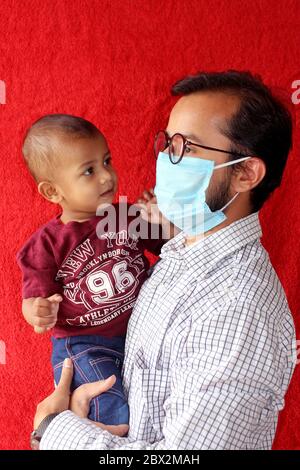 Vater trägt chirurgische Maske im Gesicht und tragen sein Baby Sohn spielen zusammen, Vatertag Konzept während Corona-Virus, covid-19 auf rotem Hintergrund Stockfoto