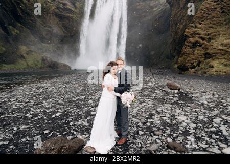 Der Bräutigam umarmt die Braut, es schneit. Hochzeitspaar in der Nähe des Skogafoss Wasserfalls. Hochzeit in Island. Stockfoto