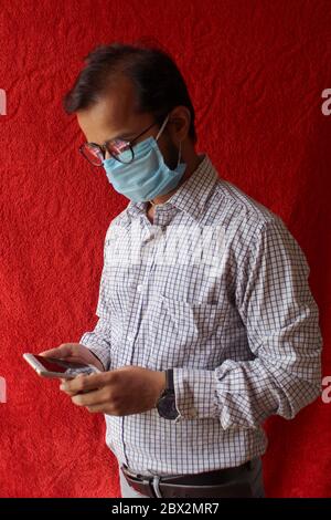 Bärtiger Erwachsener Mann mit Smartphone, während das Tragen chirurgischer Maske auf rotem Hintergrund. Gesundheit, Epidemien, soziale Medien, Kommunikation und Lifestyle-Stills Stockfoto