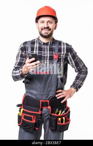 Porträt des jungen Handwerkers mit Smartphone in Schutzkleidung, Helm und Werkzeuggürtel auf weißem Hintergrund Stockfoto