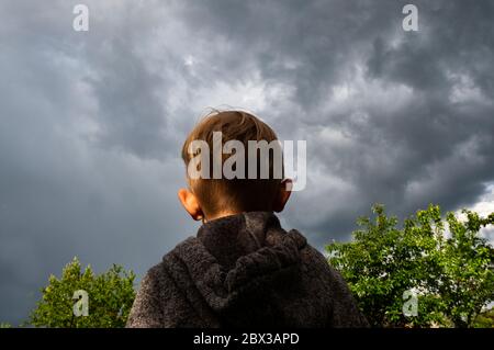 Der kleine blonde Kaukasusjunge in einer grauen Jacke mit Kapuze sieht einen stürmischen Himmel über dem Garten an Stockfoto