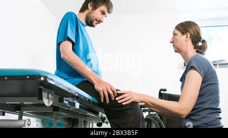 Physiotherapeut, der mit behinderten Menschen im Rollstuhl auf einem Therapietisch trainiert. Stockfoto