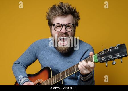 Lustige Hipster in einem alten Pullover spielt eine akustische Gitarre und singt laut, auf gelbem Hintergrund. Hobbys, Lifestyle. Stockfoto