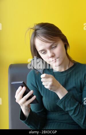 Ein junges, schönes Mädchen von europäischem Aussehen in einer blauen Jacke sitzt in einem Café mit einem Handy auf der Couch vor dem Hintergrund einer gelben Wand. Schaut durch eine Lupe auf die Gadgets-Anzeige. Stockfoto