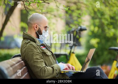 Ein Mann in einer medizinischen Gesichtsmaske, um die Ausbreitung Coronavirus zu vermeiden, arbeitet ferngesteuert auf einem Laptop im Park. Ein Mann trägt eine Pilotensonnenbrille sitzt auf einer Bank auf Stockfoto