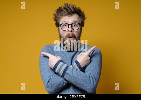 Überraschender Mann mit zerzaust Haar und Bart zeigt Finger in verschiedene Richtungen mit gekreuzten Armen und sieht verwirrt aus. Stockfoto