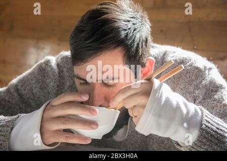 Porträt eines bärtigen Mannes, der in einer Holzhütte aus einer Schüssel sitzt und aus einer Schüssel isst, mit Essstäbchen. Stockfoto