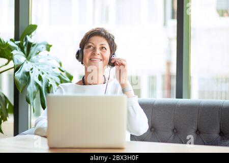 Die moderne Frau mittleren Alters tragen, halten Sie das Headset am Tisch sitzen vor dem Computer, Laptop weg schauen bis zu der Seite, glücklich lächelnd. Positive Stockfoto