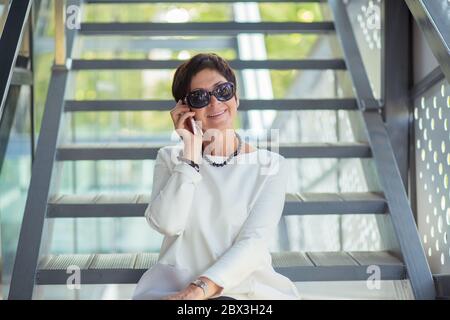 Fröhliche mittlere Alter stilvolle Frau in schwarzer Sonnenbrille sitzt auf der Treppe auf der Straße entspannend Blick auf Sie Kamera und lächelnd glücklich tragen weiß BL Stockfoto
