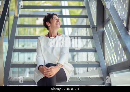 Fröhliche Frau mittleren Alters sitzt auf der Treppe auf der Straße entspannen Tagträumen Blick auf den Himmel oder Koptraum und lächelt glücklich tragen weißen Blous Stockfoto
