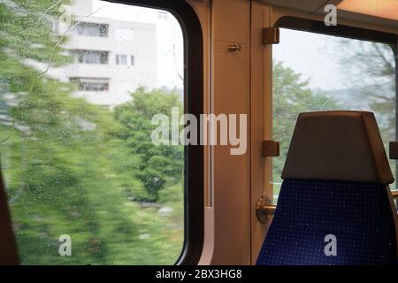 Leerer Pendlerzug an einem regnerischen Tag sind die Fenster mit Regentropfen bedeckt, die Bäume und Gebäude blinken in der Bewegung des Zuges. Stockfoto