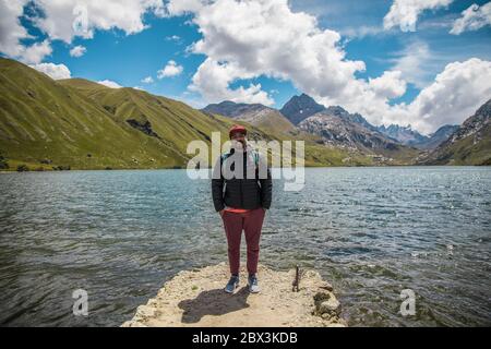 Brauner Mann, mit Jacke, Mütze und Reisetasche, in der Nähe eines Sees, Berge und blauen Himmel Stockfoto