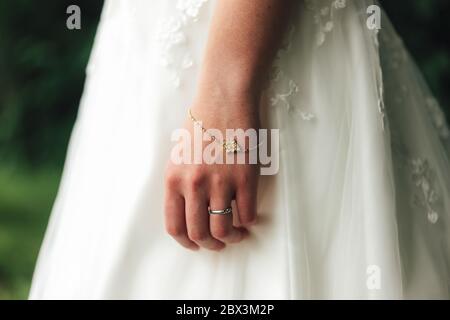 Nahaufnahme von Braut Schmuck auf ihrer Hand gegen weiße Hochzeitskleid. Goldenes Armband mit Edelsteinen und silbernem Ehering verziert. Stockfoto