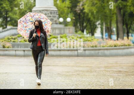 Ein maskiertes Mädchen läuft auf der Straße. Ein Mädchen in einer Schutzmaske geht im Park mit einem Regenschirm im Regen. Coronavirus-Infektion COVID-19 Stockfoto