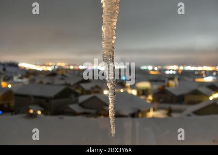 Vor einer verschwommenen Sicht auf ein Unterteilungsviertel, Stadt und Berge in Spokane, Washington, hängt im Winter ein Eiszapfen Stockfoto