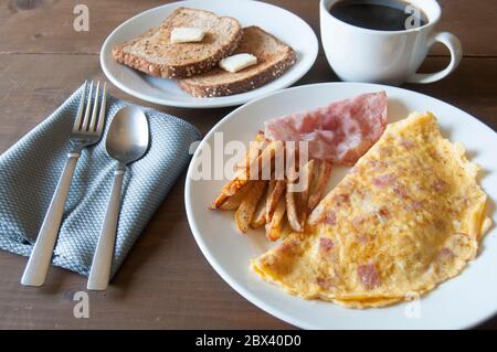 Omelette mit Pommes, Schinken und Toast mit Butter auf dem Brot, schwarzer Kaffee als Teil des Frühstücks Stockfoto