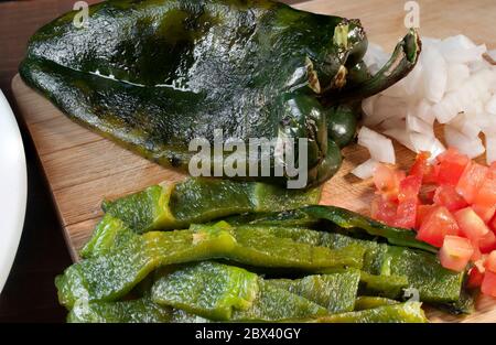 Chili poblano oder chili ancho, geschält und in Scheiben mit Zwiebeln und Tomaten auf der Seite, auf einem Holzbrett, Poblano Pfeffer oder Pasilla Pfeffer Stockfoto