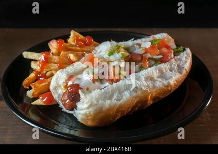 Hot Dog withonion Crean und Tomaten mit Pommes auf der Seite, auf einem schwarzen Teller. Stockfoto