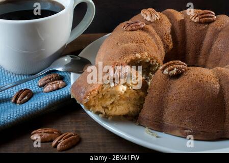 Eine Scheibe einer Nuss-ringförmigen Kuchen, auf einem weißen Teller und Nüsse auf dem Kuchen und in einer blauen Serviette mit einem weißen Becher oder Becher auf der Seite. Auf einem Holztisch Stockfoto