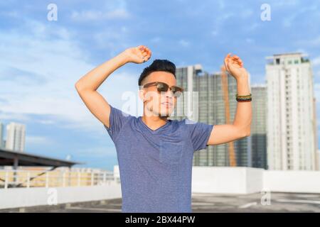 Junge schöne multi ethnischen Mann mit erhobenen Armen und trägt Sonnenbrille gegen den Blick auf die Stadt Stockfoto
