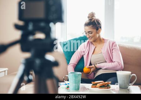 Junge Frau vlogging mit einer Tasse Tee und Sandwiches auf dem Tisch, während Brillen und hält eine Orange Stockfoto