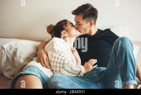 Niedliches kaukasisches Paar, das im Bett liegt und sich gegenseitig küsst, Jeans und weißen Strickpullover trägt Stockfoto
