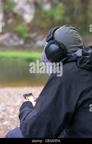 Stylischer junger Mann oder Hipster, der Musik in Kopfhörern hört und Smartphone in der Hand sitzt am Strand. Musik-Lounge, entspannend. Blick von hinten. Befreit Stockfoto