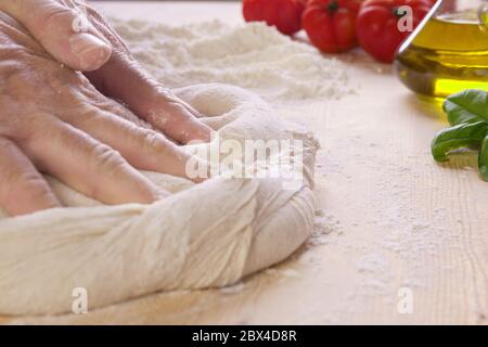 Makro Nahaufnahme von Mann Chef Hände kneten Pizzateig auf Holzplatte und Mehl, extra natives Öl, Tomaten und Basilikum im Hintergrund Stockfoto