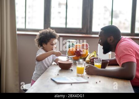 afroamerikanischer Mann in einem rosa T-Shirt unterhaltsam seine Tochter, während sie essen Stockfoto