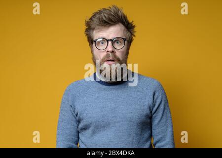 Aufgeregt zotteligen Mann mit Brille und einem alten Pullover schaut auf die Kamera mit einem schockierten Ausdruck im Gesicht, auf einem gelben Hintergrund. Stockfoto