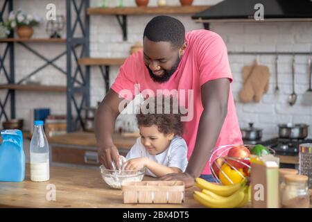 afroamerikanischer bärtiger Mann spielt mit seiner Tochter, die das Mehl in der Schüssel rührt Stockfoto
