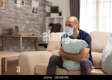 Ängstlicher Mann hält fest ein Kissen während der Selbstisolierung trägt Einwegmaske. Stockfoto