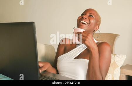 Junge schöne und glückliche schwarze Afro amerikanische Frau lächelnd aufgeregt Spaß im Internet mit sozialen Medien auf Laptop-Computer zu Hause Wohnzimmer c Stockfoto
