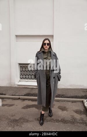 Junge stilvolle Brünette Frau trägt lange graue Mantel, Anzug und Schuhe, zu Fuß entlang der Straßen, attraktive Frau in trendigen Casual Outfit und Sonnenbrille Stockfoto