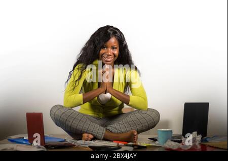 Junge glückliche und attraktive afroamerikanische Geschäftsfrau tun Yoga sitzen im Büro unordentlich Schreibtisch voller Papierkram lächelnd entspannt und sorglos in einem Stockfoto