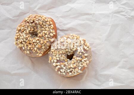 Braun glasierte Donuts mit Nüssen auf zerknittertem beigefarbenem Papierhintergrund bestreut. Ungesunde kalorienreiche Lebensmittel. Leere Stelle für Text. Kopierbereich Stockfoto