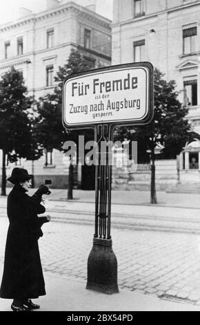 Eine Frau mit Hund im Arm steht auf einer Straße vor dem Augsburger Bahnhof vor einem Schild mit der Aufschrift "der Einzug nach Augsburg ist für Fremde gesperrt". Aufgrund des vorherrschenden Wohnungsmangels widersetzten sich viele große Stadtgemeinden nach dem Ersten Weltkrieg der weiteren Einwanderung von außerhalb der Stadt. Das Warnschild soll Neuankömmlinge abschrecken. Stockfoto