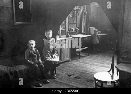 Zwei Kinder sitzen auf einem Bett in einem dunklen Haus. Rechts davon steht die Küche, in der ein anderes (verschwommenes) Kind steht. Die Küche und der Schlafbereich sind durch Vorhänge getrennt. Aufgrund der französischen Besetzung kam es im Rhein-Ruhr-Gebiet zu Wohnungsmangel. Stockfoto