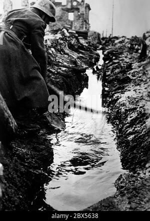 Ein deutscher Soldat versucht, mit einer Schaufel Wasser aus einem Graben zu entfernen. Im Hintergrund befindet sich ein Haus, das durch Bombenangriffe zerstört wurde. Während der Einkreisungsschlacht von Kholm verteidigte die Kampfgruppe Scherer die Stadt erfolgreich für 105 Tage. (Ein Foto der Propaganda Company (PK) des Kriegsberichterstatters Richard Muck, der Anfang März in die Tasche flog). Stockfoto