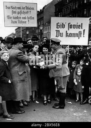 Während einer von der Deutschen Arbeitsfront organisierten Straßenversammlung des Winterhilfswerks des Deutschen Volkes werden nach der Münchner Konferenz Friedenslogans gehalten (Reich und Frieden sind gesichert!). Wir danken und helfen dem Führer). Während der Spendenaktion werden 'Fuhrerbüchlein' für 'soviel man sich leisten kann' (Plakat) verkauft. Stockfoto