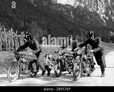 Drei Mitglieder der Motor Hitler Jugend schieben ihre Motorräder auf einer Landstraße für eine Übungsfahrt. Links ist eine Gruppe, die sie beobachtet. Im Hintergrund sind Berge. Das Motorrad in der Mitte ist ein Puch 200. Links und rechts sind Puch 125 T Motorräder. Stockfoto