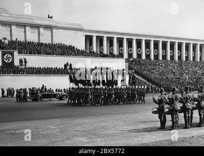 Blick auf den formationsmarsch des Reichsarbeitsdienstes mit Fahnen und Pik auf dem Zeppelinfeld vorbei an Adolf Hitler in seinem Mercedes und der Tribüne des Zeppelinfelds. Links vor Hitler stehen Konstantin Hierl und Rudolf Hess. Auf der Tribüne steht ein Kamerateam. Im Vordergrund eine Drummergruppe des Rad. Stockfoto